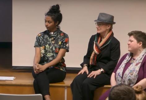 Two artists, a young, dark haired woman in a brightly colored patterned blouse and an older woman with short white hair in a blazer and fedora. To the far right is a curator.