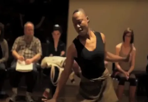 Image of Nora Chipaumire dancing. She is a Black woman with a shaved head dancing in front of a seated audience in a museum gallery. She wears a black leotard and brown skirt.