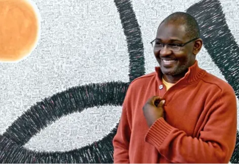 Image of a smiling Nigerian man, the artist Victor Ekpuk, in front of a painting that combines abstraction and nsibidi, a form of script used in his home country.