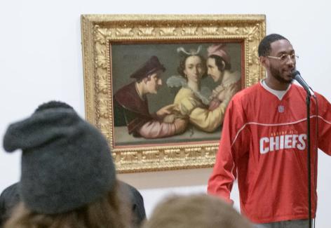 A musician stands in front of a painting and is performing a rap in front of a crowd.