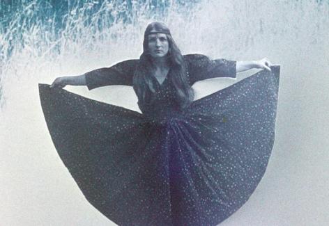 Screenshot of a frame from the Bea Nettles talk at Visual Studies Workshop in Rochester, NY in 1974. It shows a self portrait of the artist in a star covered dress, holding her skirt out to the sides. It is hand-colored in blue tones like night and sky.