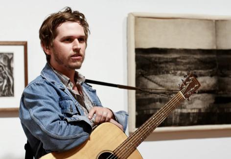 A man with an acoustic guitar stands next to an artwork made of printed and stuffed fabric.