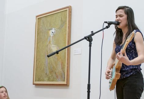 A musician plays an electric guitar in front of a painting. She is playing to a crowd seated on the floor.