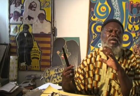 Senegalese artist Yelimane Fall in a skullcap and brown and yellow patterned caftan sitting in his studio talking to the camera. In the background is colorful art. Fall has a beard and brown skin.