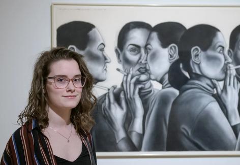 A young women stands in front of a large drawing featuring the artist in different poses, sometimes smoking a cigarette.